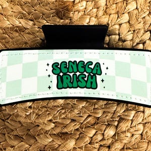 K&ouml;nnte beinhalten: Ein gr&uuml;ner und wei&szlig;er karierter Haarklammer mit dem Text "SENECA IRISH" in gr&uuml;nen Buchstaben.