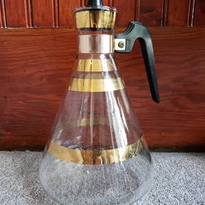 May include: A clear glass pitcher with a black handle and gold stripes. The pitcher has a black lid.