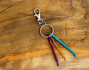 Pode incluir: Um chaveiro prateado com um anel e três agulhas de crochê coloridas: azul-turquesa, dourado e roxo.