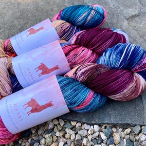 May include: Three skeins of hand-dyed yarn featuring variegated colors of deep magenta, bright pink, and teal blue. Each skein is wrapped in a white paper label with a red llama silhouette and the text HANKS yarning company luxury yarn.