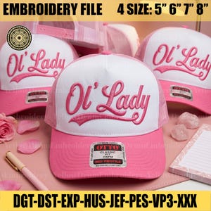 May include: A white and pink trucker hat featuring the embroidered text Ol' Lady in pink script on the front panel. The hat includes a pink brim, a white foam front, and a pink mesh back. A sticker on the brim reads OTTO CLASSIC FIT OSFM.