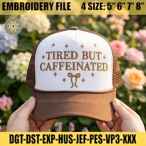 May include: A brown and white trucker hat with the text "TIRED BUT CAFFEINATED" embroidered on the front. The hat has a curved brim and a mesh back. The hat is being held up by a person.