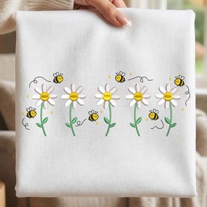 May include: White fabric with an embroidered design of daisies and bees. The daisies have white petals and yellow centers. The bees are yellow and black. The stems and leaves are green. The design is centered on the fabric.