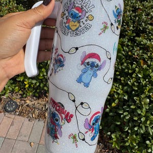 Pu&ograve; includere: Un bicchiere bianco glitterato con Stitch con un cappello da Babbo Natale, con la scritta "Official Cookie Tester" e "Stitchmas". Il design include luci a stringa e bastoncini di zucchero. Il bicchiere ha un manico bianco.