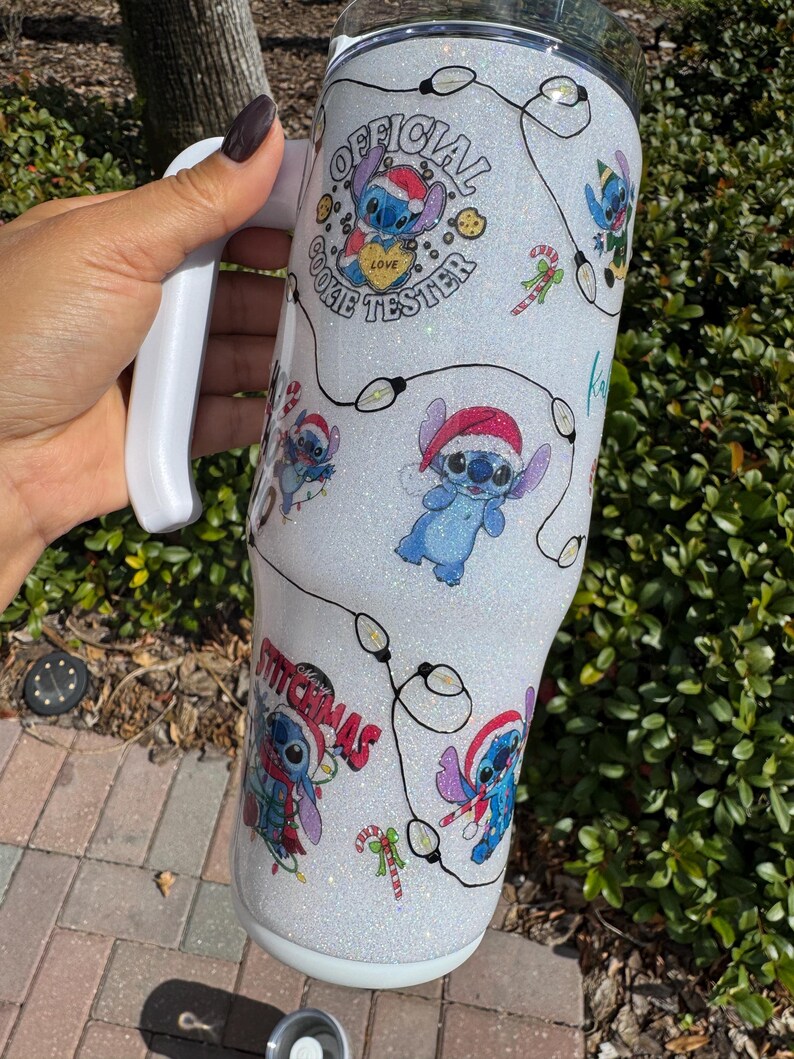 Pu&ograve; includere: Un bicchiere bianco glitterato con Stitch con un cappello da Babbo Natale, con la scritta "Official Cookie Tester" e "Stitchmas". Il design include luci a stringa e bastoncini di zucchero. Il bicchiere ha un manico bianco.