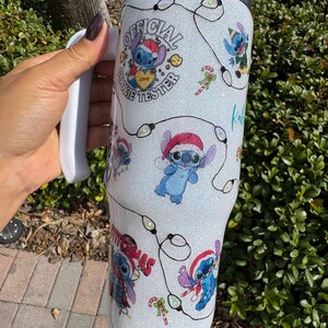 Pu&ograve; includere: Un bicchiere bianco glitterato con personaggi dei cartoni animati di Stitch con cappelli da Babbo Natale e luci natalizie. Il bicchiere ha il testo "OFFICIAL COOKIE TESTER" e "MERRY CHRISTMAS". Include un manico bianco.