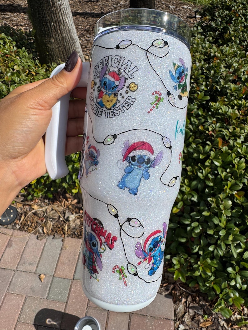 Pu&ograve; includere: Un bicchiere bianco glitterato con personaggi dei cartoni animati di Stitch con cappelli da Babbo Natale e luci natalizie. Il bicchiere ha il testo "OFFICIAL COOKIE TESTER" e "MERRY CHRISTMAS". Include un manico bianco.