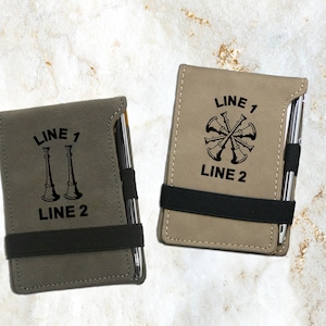 Pode incluir: Dois cadernos de couro sintético personalizados com uma caneta. Um caderno tem uma ilustração em preto e branco de mangueiras de incêndio cruzadas. O outro caderno tem uma ilustração em preto e branco de dois chifres de fogo. Ambos os cadernos têm o texto "LINE 1" e "LINE 2" neles.