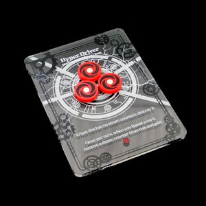 May include: A clear plastic card game card with a black and white design featuring gears and lightning bolts. The card has the text "Hyper Driver" and game instructions. Three red plastic tokens are on the card.