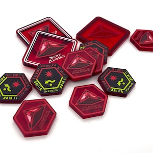 May include: A collection of red and black game tokens with geometric designs. The tokens include square, hexagonal, and triangular shapes. Some tokens feature a question mark and star symbol in yellow and green. Other tokens have a triangular design with text.