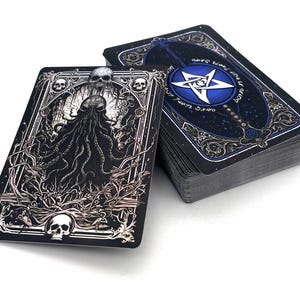 Puede incluir: Una baraja de cartas del tarot con un diseño oscuro y gótico. La carta frontal presenta una detallada ilustración en blanco y negro de una figura con cabeza de calavera y tentáculos. Los dorsos de las cartas son de color azul oscuro con un pentagrama blanco y detalles florales.