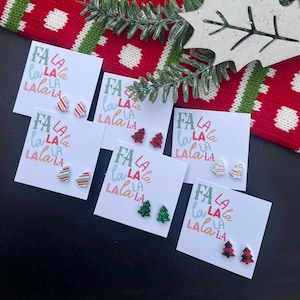 May include: Five cards with the text "FA LA LA LA LA" printed on them. Each card has a pair of small Christmas tree earrings with glitter. The earrings are red, green, and white.