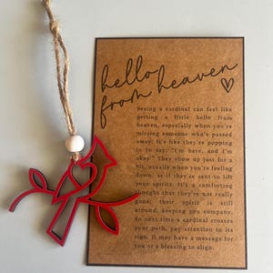 Op de afbeelding: Een rood houten ornament van een kardinaalvogel met een hart op zijn borst, zittend op een tak. Het ornament hangt aan een touwtje van touw. De kaart heeft de tekst "hello from heaven" met een hartje. De tekst op de kaart beschrijft de kardinaal als een teken van geliefden die zijn overleden.