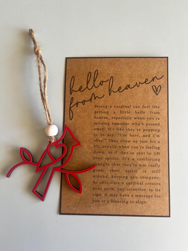 Op de afbeelding: Een rood houten ornament van een kardinaalvogel met een hart op zijn borst, zittend op een tak. Het ornament hangt aan een touwtje van touw. De kaart heeft de tekst "hello from heaven" met een hartje. De tekst op de kaart beschrijft de kardinaal als een teken van geliefden die zijn overleden.