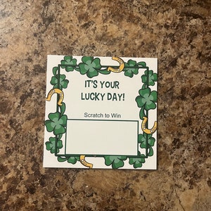 May include: A white scratch-off card with the text "IT'S YOUR LUCKY DAY!" and "Scratch to Win". The card is decorated with green shamrocks and gold horseshoes. The card has a blank area to scratch and reveal a prize.