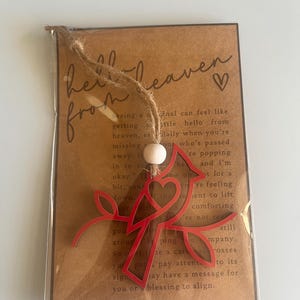 Op de afbeelding: Een rood metalen kardinaal ornament met een hartvormige uitsnijding. Het ornament is bevestigd aan een bruine kaart met de tekst "hello from heaven" en een klein hart.