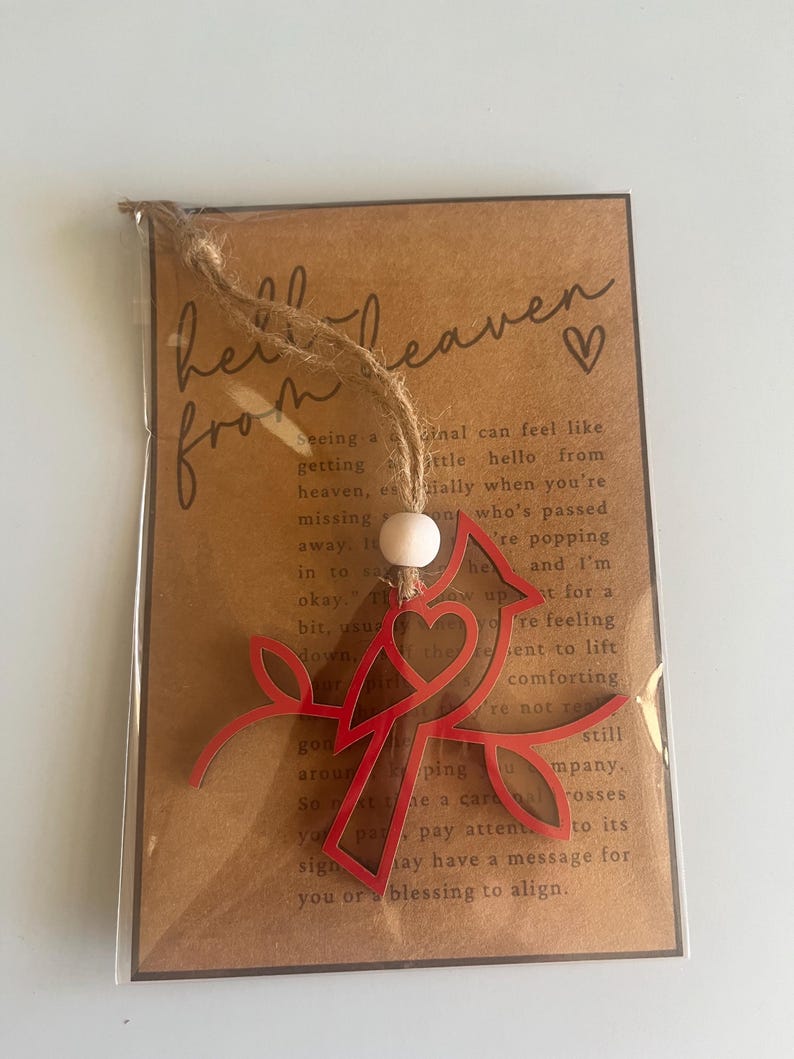 Op de afbeelding: Een rood metalen kardinaal ornament met een hartvormige uitsnijding. Het ornament is bevestigd aan een bruine kaart met de tekst "hello from heaven" en een klein hart.