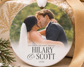 Peut inclure: Une décoration de Noël ronde en céramique blanche avec une photographie d'un couple qui s'embrasse. Le texte indique OUR FIRST CHRISTMAS MARRIED HILARY & SCOTT 2025. La décoration est suspendue par un ruban blanc sur un cadeau.