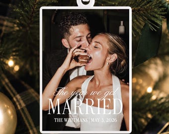 Peut inclure: Un ornement rectangulaire blanc présentant une photographie d'un couple mangeant un gâteau. Le texte indique : The year we got MARRIED THE WHITMANS | MAY 3, 2026. L'ornement est suspendu par un ruban blanc devant un sapin vert.