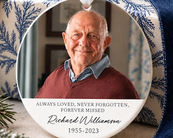 May include: A round white ceramic memorial ornament featuring a portrait of an older man in a maroon sweater. The text reads: ALWAYS LOVED, NEVER FORGOTTEN, FOREVER MISSED, Richard Williamson, 1955-2023. It hangs from a white ribbon.