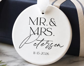 May include: A round white ceramic ornament featuring black text that reads MR. & MRS. Petersen and the date 11-15-2026. The ornament has a small hole at the top with a white ribbon attached for hanging, displaying a clean and smooth surface.
