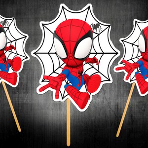 May include: Three Spiderman character cutouts on wooden sticks. Each cutout features a red and blue Spiderman figure with a white spiderweb background and a small black spider. The background is a dark grey.