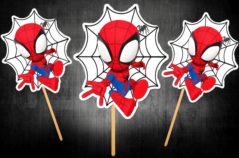 May include: Three Spiderman character cutouts on wooden sticks. Each cutout features a red and blue Spiderman figure with a white spiderweb background and a small black spider. The background is a dark grey.