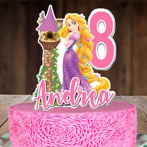 May include: A pink frosted cake with a decorative floral pattern, topped with a Rapunzel cake topper. The topper features a cartoon illustration of Rapunzel, a tower, the number 8, and the name "Andrea" in pink script.