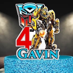 Puede incluir: Un adorno para tarta con un personaje de robot amarillo y negro de Transformers, con el número 4 en rojo y el nombre "Gavin" en azul. La tarta está decorada con glaseado azul.
