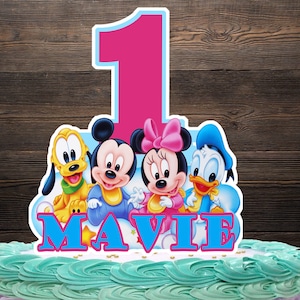 Puede incluir: Un adorno para tarta con Mickey Mouse, Minnie Mouse, Pluto y el Pato Donald. El número 1 se muestra en rosa con un contorno azul claro. El nombre "MAVIE" está escrito en letras azules. La tarta tiene glaseado turquesa.