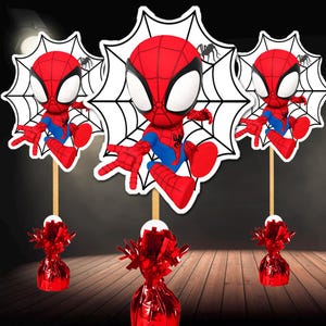 May include: Three Spider-Man party decorations. Each features a red and blue Spider-Man figure on a wooden stick, set against a white spiderweb backdrop. Red foil balloon weights are at the base of each decoration.