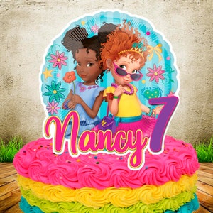 Op de afbeelding: Een kleurrijke verjaardagstaart met regenboogglazuur, bekroond met een cartoonafbeelding van twee meisjes, de naam "Nancy" en het nummer "7". De taart heeft lagen roze, gele en blauwe glazuur.