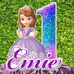 May include: A birthday cake topper featuring a cartoon princess in a purple dress and a number one. The princess has brown hair and a tiara. The name "Emie" is written in purple script. The number one is decorated with blue and purple glitter.
