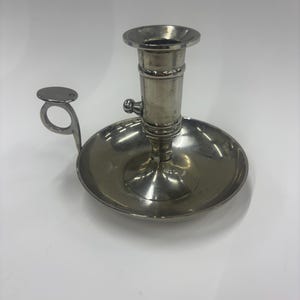 Puede incluir: Un candelero plateado con base redonda y copa cilíndrica para vela. Cuenta con un pequeño asa con un disco circular. El candelero es de metal y tiene una superficie reflectante.