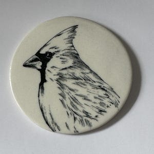 K&ouml;nnte beinhalten: Ein wei&szlig;er Keramikuntersetzer mit einer schwarzen Linienzeichnung eines Kardinals.