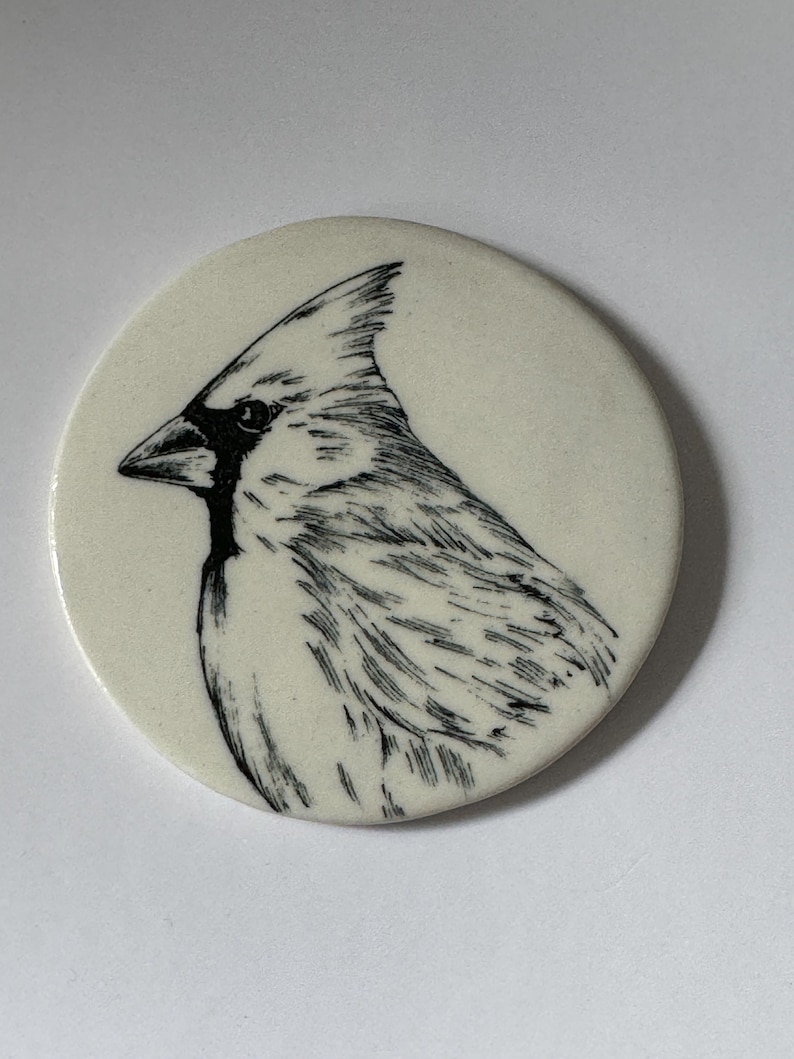 K&ouml;nnte beinhalten: Ein wei&szlig;er Keramikuntersetzer mit einer schwarzen Linienzeichnung eines Kardinals.