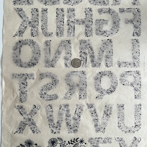 Peut inclure: Alphabet floral noir et blanc et formes de cœur imprimées sur un tissu beige. Chaque lettre est remplie de motifs floraux détaillés. Une pièce de monnaie est incluse pour la référence de taille.