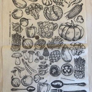 May include: A beige kitchen towel with a black and white illustration of various vegetables, fruits, and mushrooms. The illustration includes asparagus, garlic, eggplant, peppers, mushrooms, cauliflower, artichokes, corn, onions, tomatoes, and a pumpkin. The text "www.creativemarket.com" is printed in the bottom right corner of the towel.