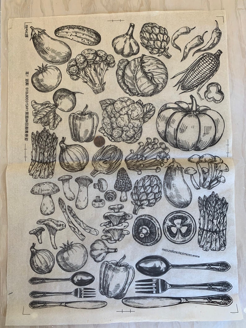 May include: A beige kitchen towel with a black and white illustration of various vegetables, fruits, and mushrooms. The illustration includes asparagus, garlic, eggplant, peppers, mushrooms, cauliflower, artichokes, corn, onions, tomatoes, and a pumpkin. The text "www.creativemarket.com" is printed in the bottom right corner of the towel.