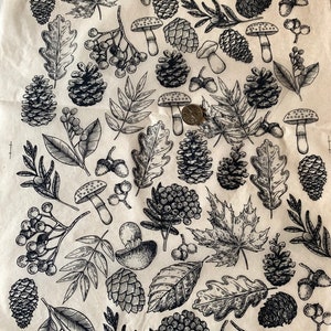 May include: A black and white illustration of a repeating pattern of autumn leaves, mushrooms, and pine cones. The pattern is on a light brown background.