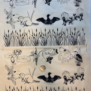 May include: A sheet of black and white rubber stamps featuring images of a frog, a bass fish, a dragonfly, a beaver, a loon, a heron, a lily pad, and two chairs. The stamps are on a beige background.