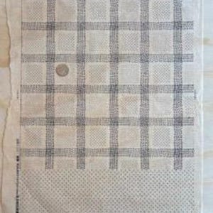 Puede incluir: Un textil rectangular con un patrón de cuadrícula en tonos grises y blancos. Una pequeña moneda descansa sobre la tela. La sección inferior presenta un diseño punteado. El textil se muestra sobre una superficie de madera clara.