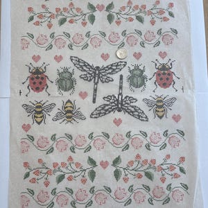 May include: A cross-stitch pattern printed on paper featuring rows of pink flowers, green leaves, and red berries. The center displays insects including ladybugs, dragonflies, and bees, interspersed with small red hearts and a US quarter coin.