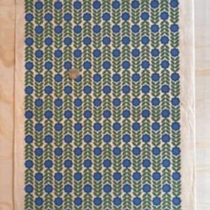 May include: A rectangular piece of fabric featuring a repeating pattern of blue flowers and green leaves against a cream backdrop. The design is symmetrical and covers the entire surface. A small, round object rests on the fabric.