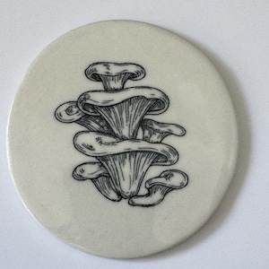 May include: A white ceramic coaster with a black and white line drawing of a cluster of mushrooms.