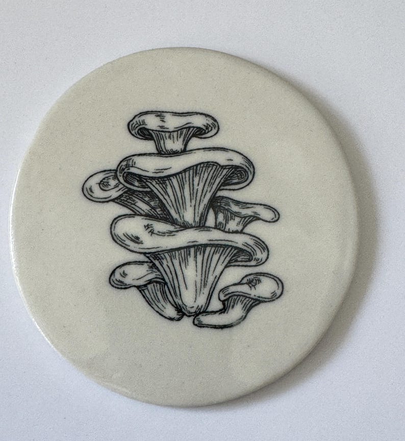 May include: A white ceramic coaster with a black and white line drawing of a cluster of mushrooms.