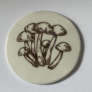 May include: A white ceramic coaster with a brown line drawing of a cluster of mushrooms.