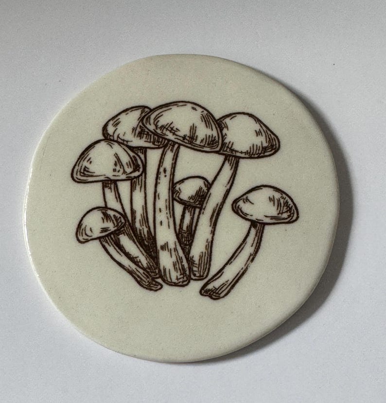 May include: A white ceramic coaster with a brown line drawing of a cluster of mushrooms.
