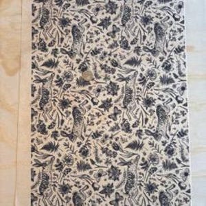 Puede incluir: Una impresión rectangular con un patrón repetido de liebres, flores y follaje azul oscuro sobre un fondo crema. El diseño incluye mariposas y otros elementos naturales. La impresión está sobre una superficie clara.