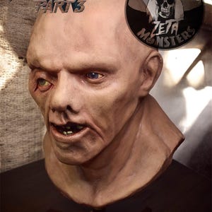 Puede incluir: Un busto realista de un personaje calvo de piel pálida con ojos asimétricos, boca ligeramente abierta que muestra los dientes y tez texturizada. El texto FRIDAY THE 13TH PART 3 aparece arriba a la izquierda, junto a un logo de ZETA MONSTERS y una etiqueta de calabaza.